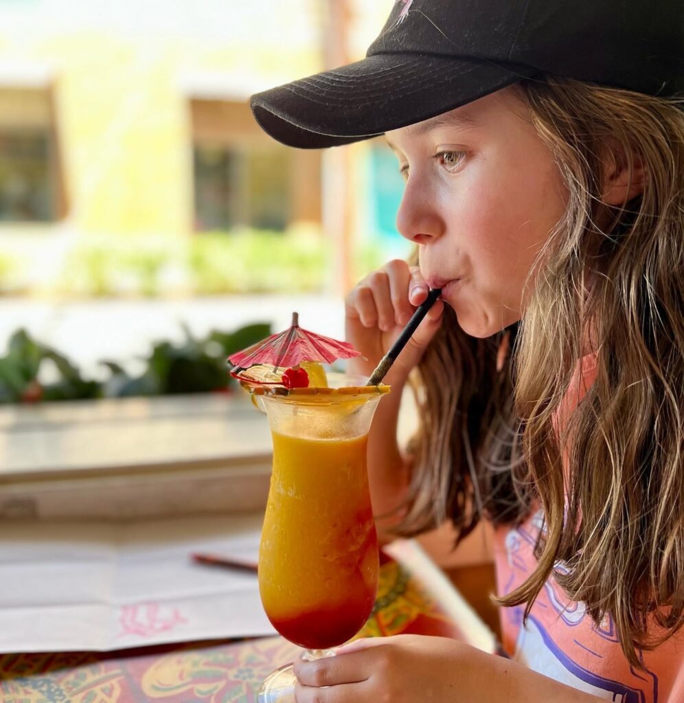 A person in a black cap sips a layered orange smoothie drink with a tiny red paper umbrella. The setting appears to be a sunny café table with a patterned tablecloth.