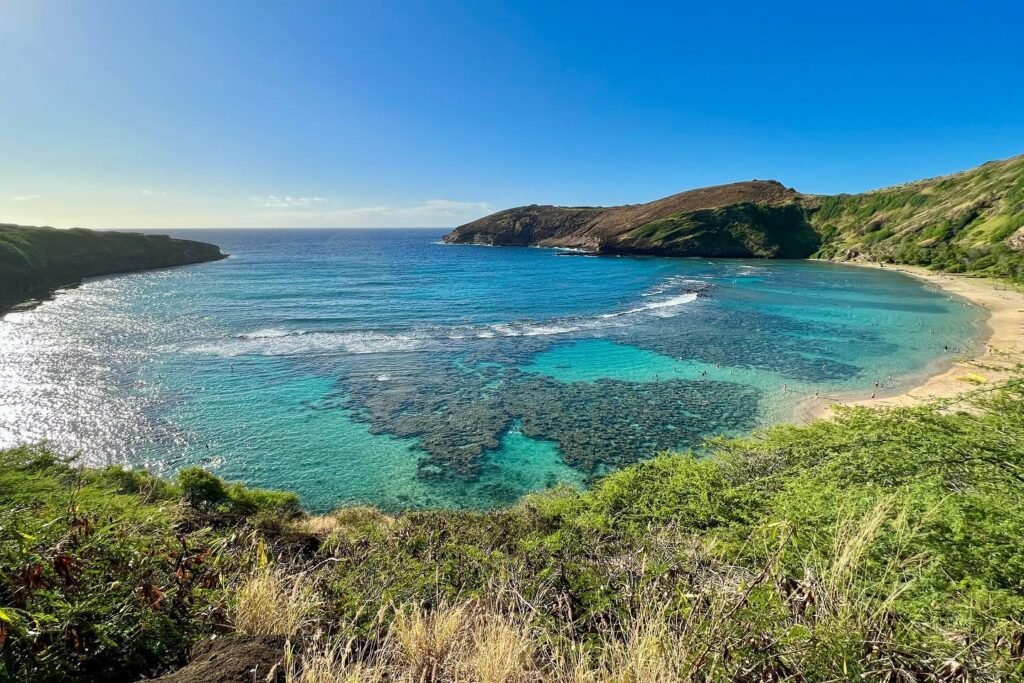 A scenic coastal landscape featuring clear turquoise waters, secluded sandy beaches, and hilly green coastlines under a bright blue sky.