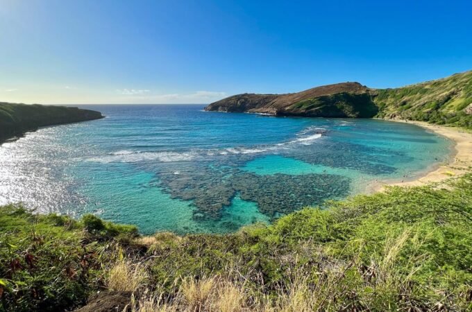 A scenic coastal landscape featuring clear turquoise waters, secluded sandy beaches, and hilly green coastlines under a bright blue sky.