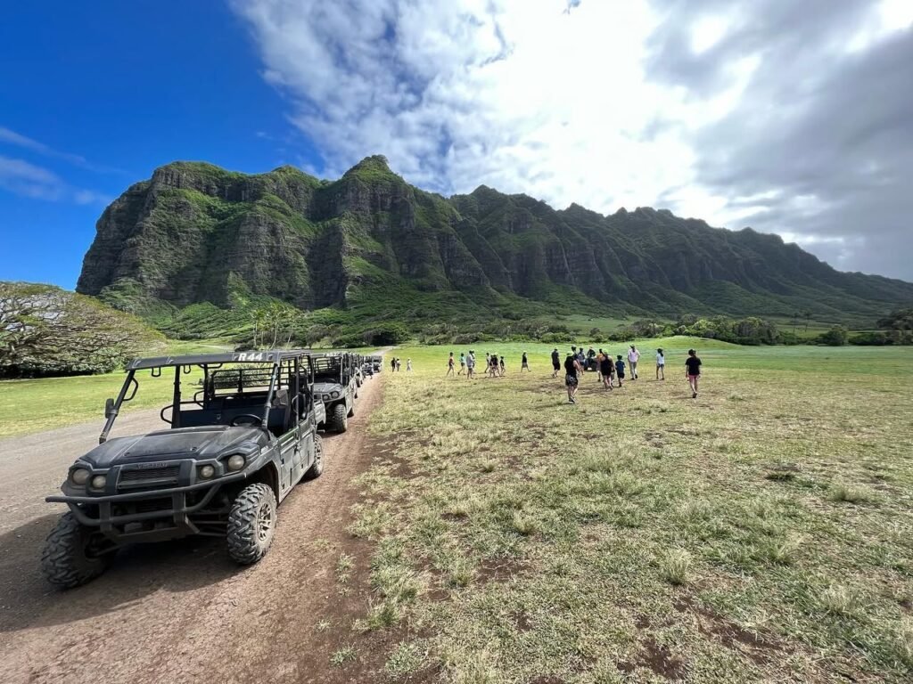 A line of ATVs drives on a dirt path beside a grassy field with hikers and a long green mountain in the background.