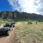 Diamond Head A line of ATVs drives on a dirt path beside a grassy field with hikers and a long green mountain in the background.