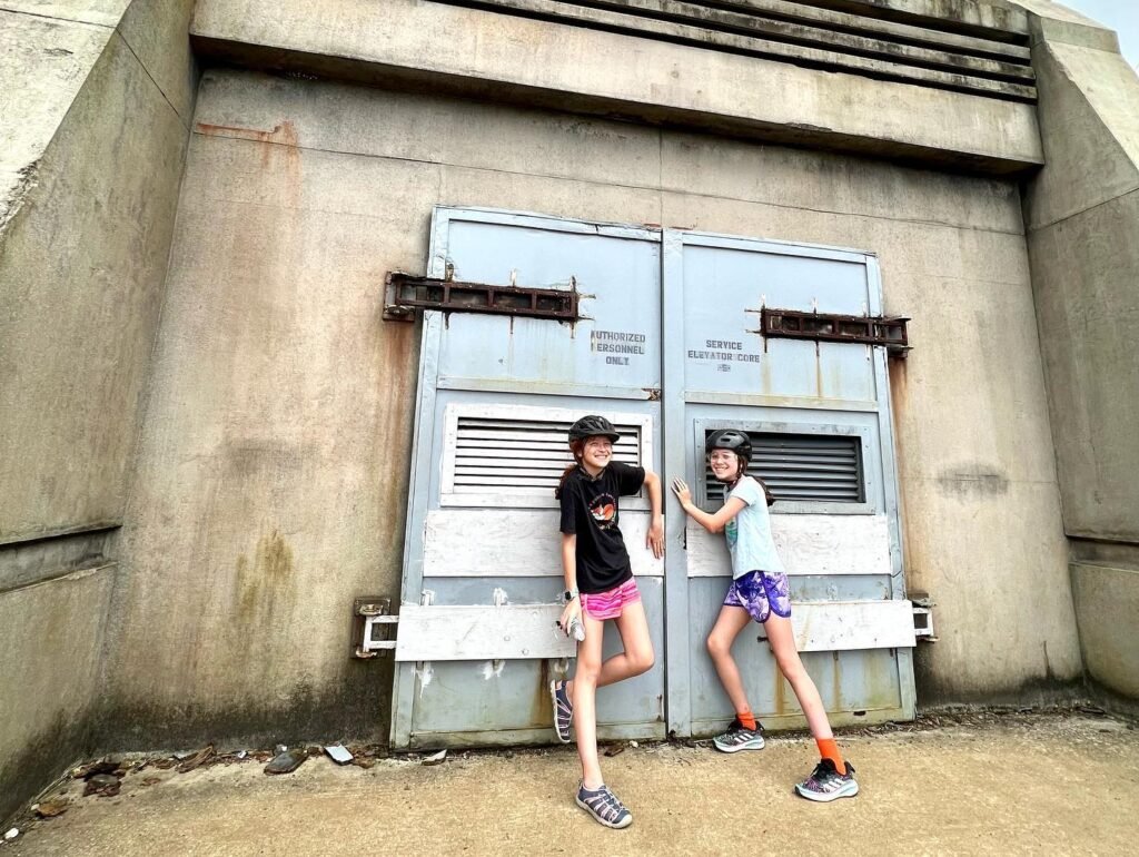 Two hikers wear protective helmets while navigating an industrial concrete structure. They stand on a dusty floor with large gray metal fixtures nearby.