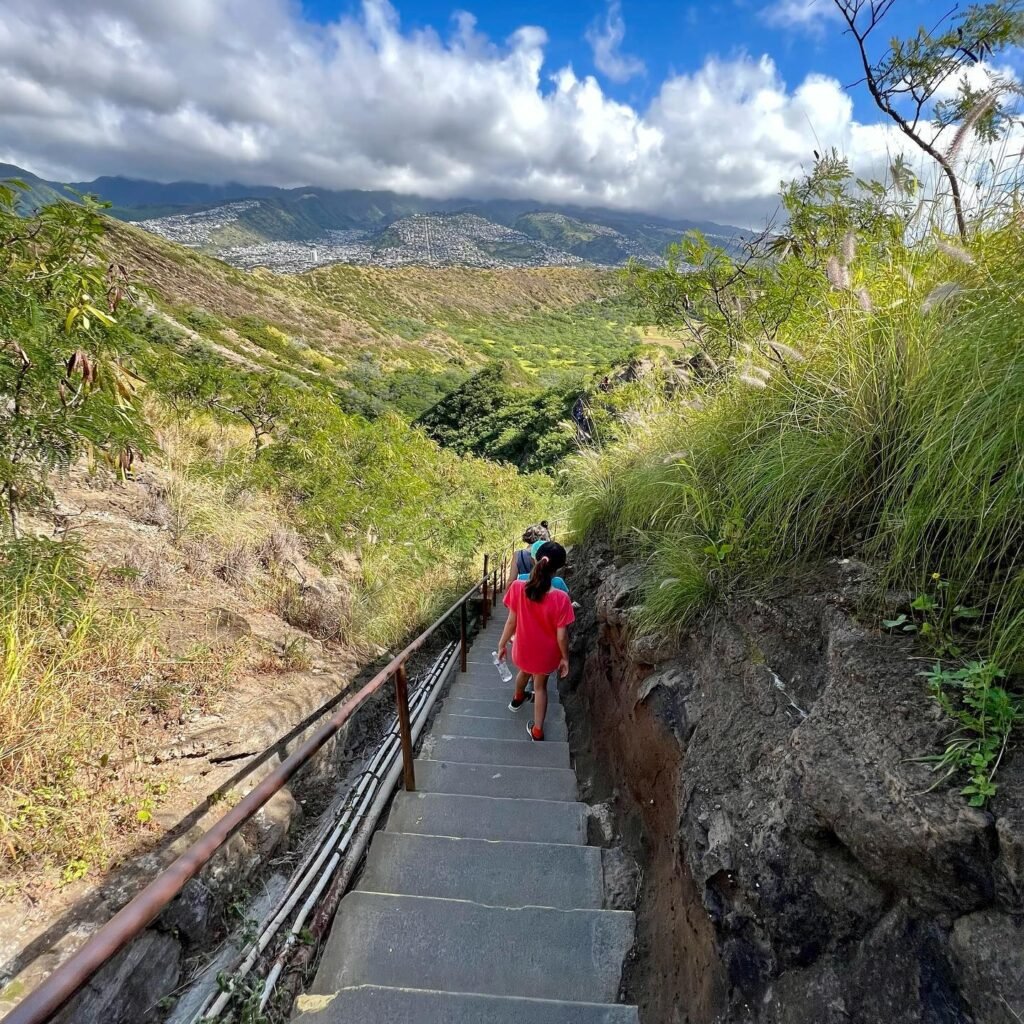 A person descends a stone staircase on the Mauna Kea Eruptions Trail, overlooking lush greenery and distant mountains. A panoramic view showcases the dramatic landscape with a cloudy sky and expansive valley below.