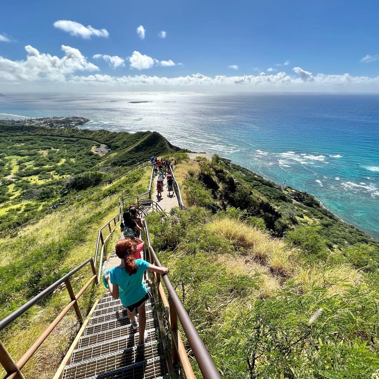 Diamond Head Hikers ascending grassy cliffs to a coastal viewpoint in Hawaii with turquoise ocean views.