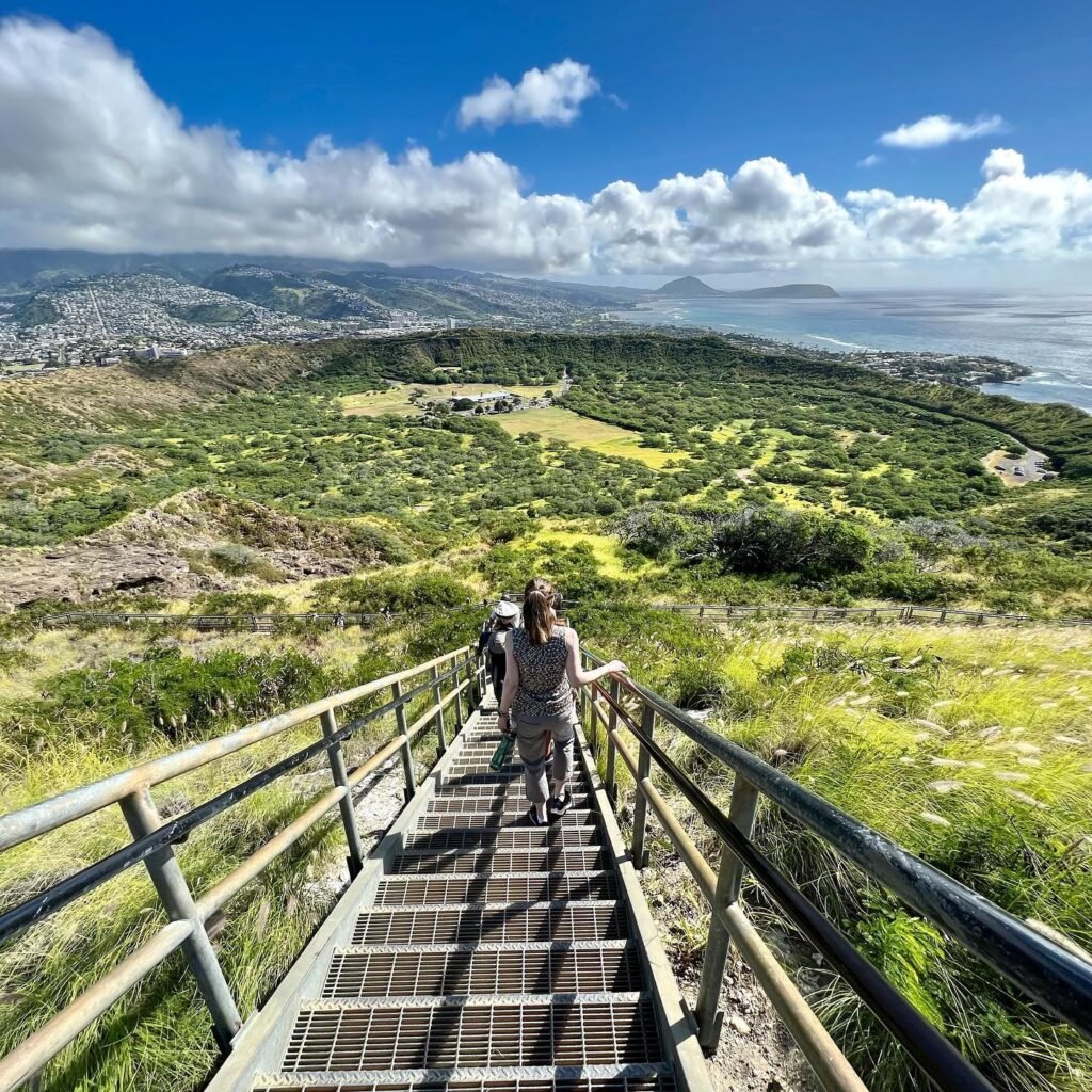 Hikers ascending a metal staircase on a mountain trail, overlooking a sweeping view of the coast and a distant city.