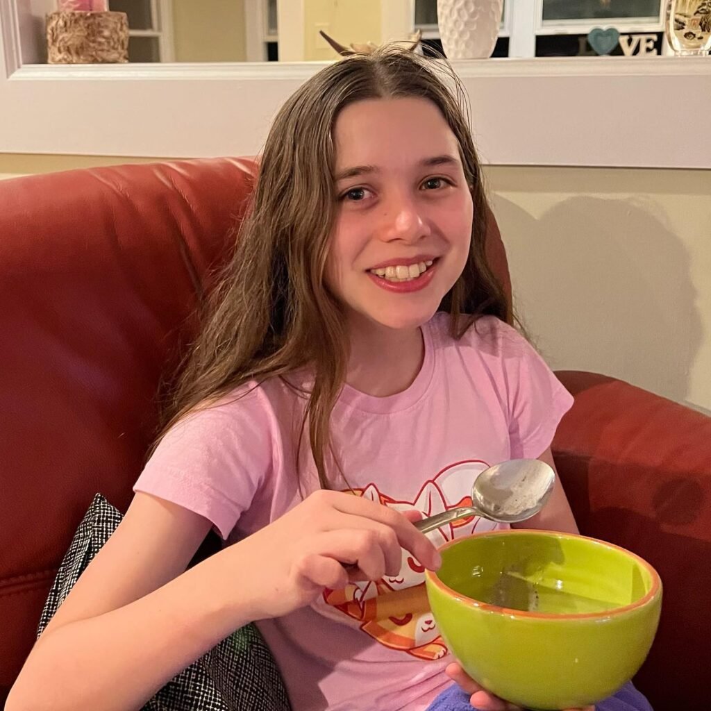 A smiling child in a pink shirt sitting on a red sofa holding a green bowl and spoon, with a cozy background.