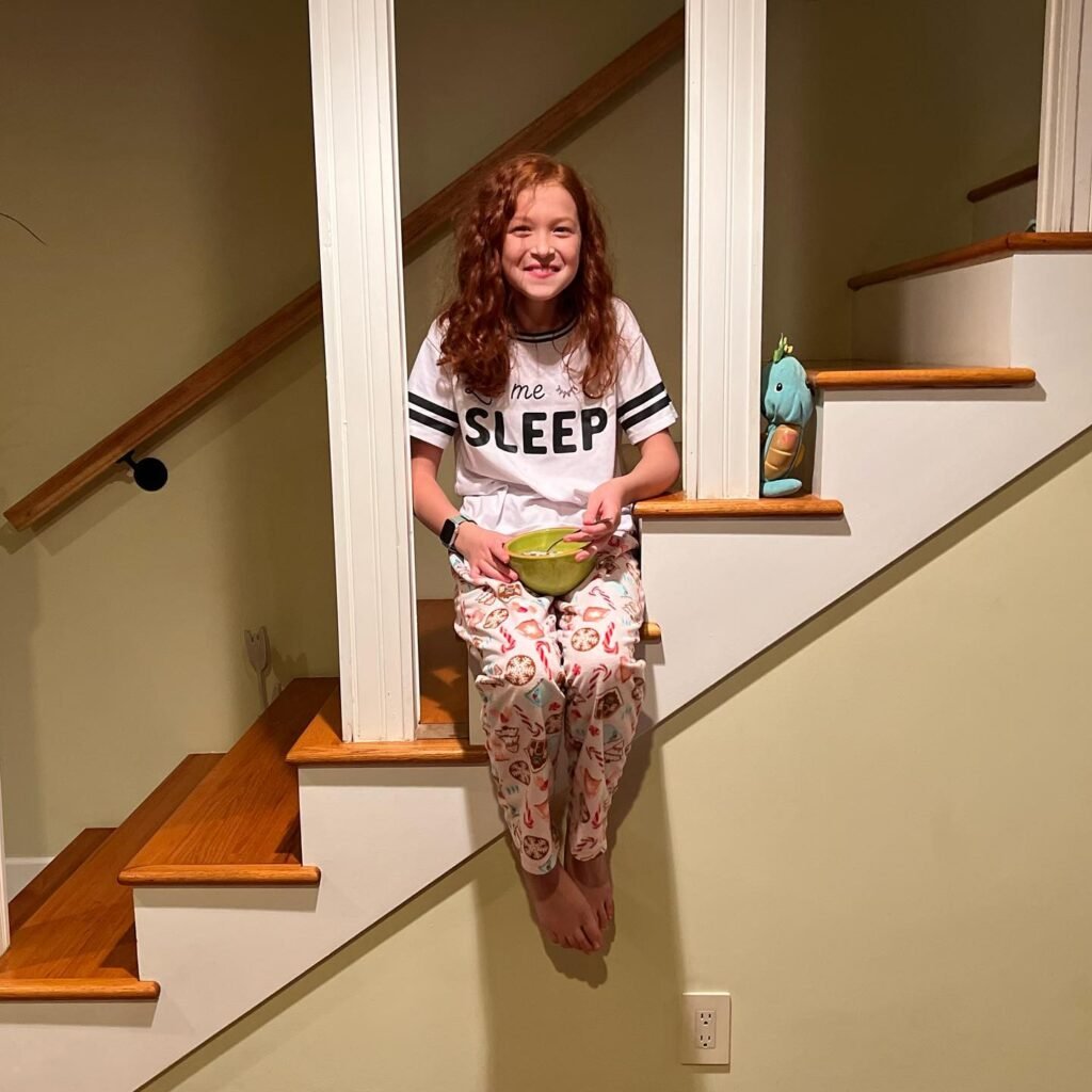 A smiling child with curly hair sits on a wooden stair landing, holding a green bowl.