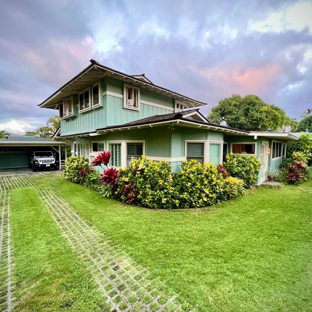 A charming two-story house with a green lawn, colorful flowers, and a carport under a cloudy sky.