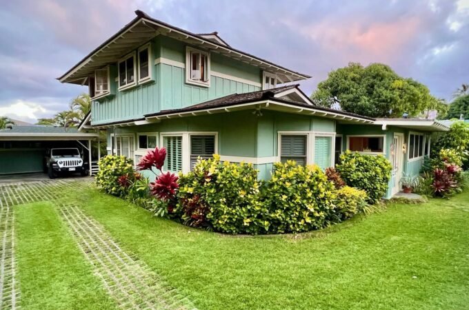 A charming two-story house with a green lawn, colorful flowers, and a carport under a cloudy sky.