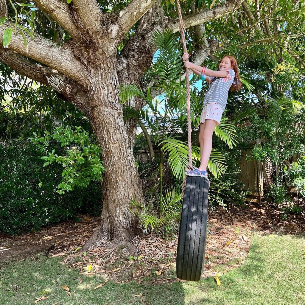 A person enjoys a homemade tire swing hanging from a lush tree in a suburban backyard.