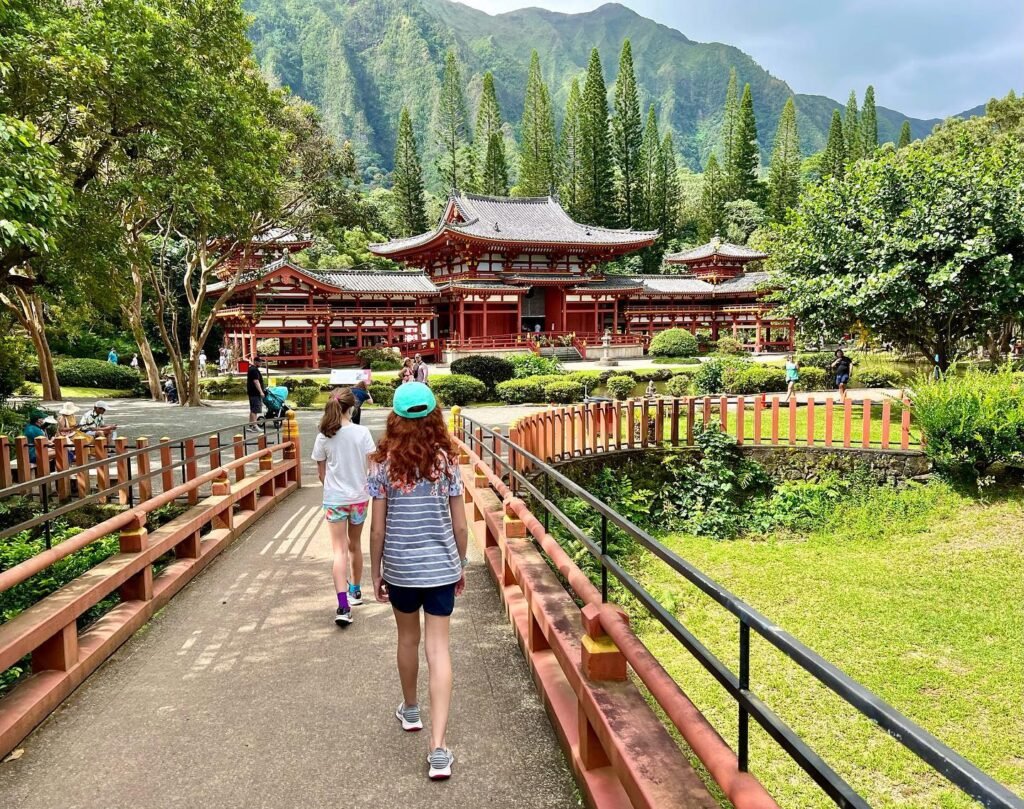 Visitors walk across a wooden bridge toward a traditional Japanese temple complex amidst lush green mountains and trees.
