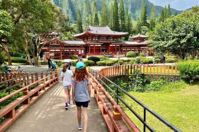 Visitors walk across a wooden bridge toward a traditional Japanese temple complex amidst lush green mountains and trees.