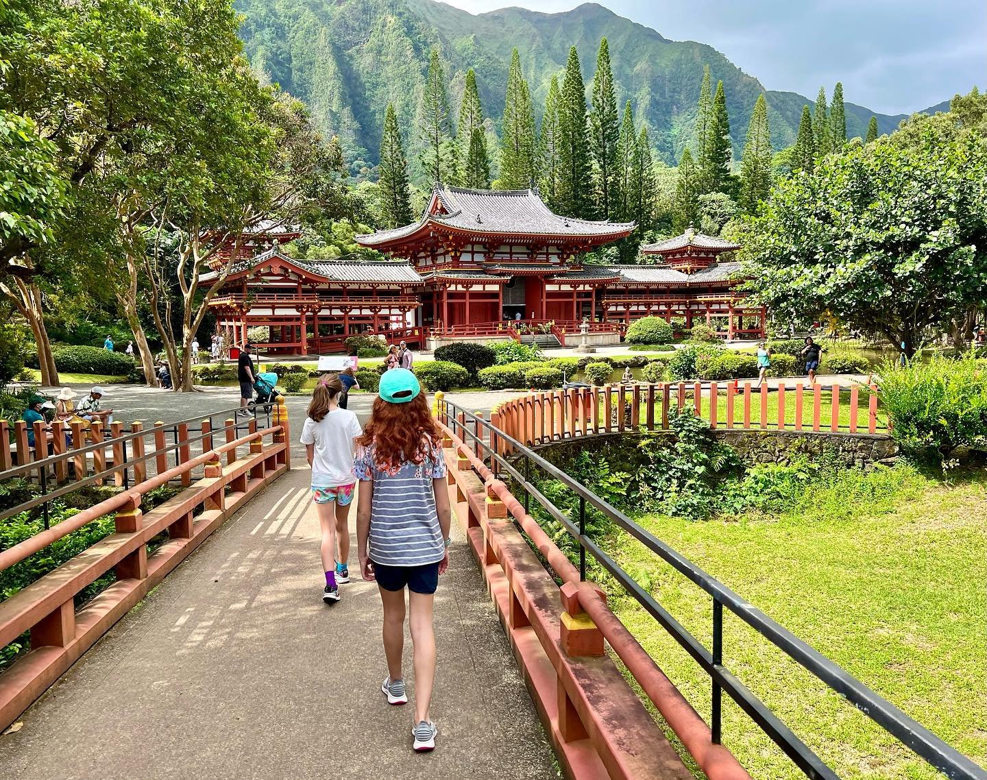 Byodo-In Temple Visitors walk across a wooden bridge toward a traditional Japanese temple complex amidst lush green mountains and trees.