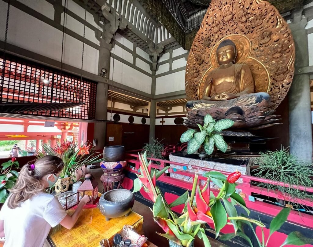 A golden Buddha statue inside a traditional Japanese temple with vibrant floral arrangements and architectural details.