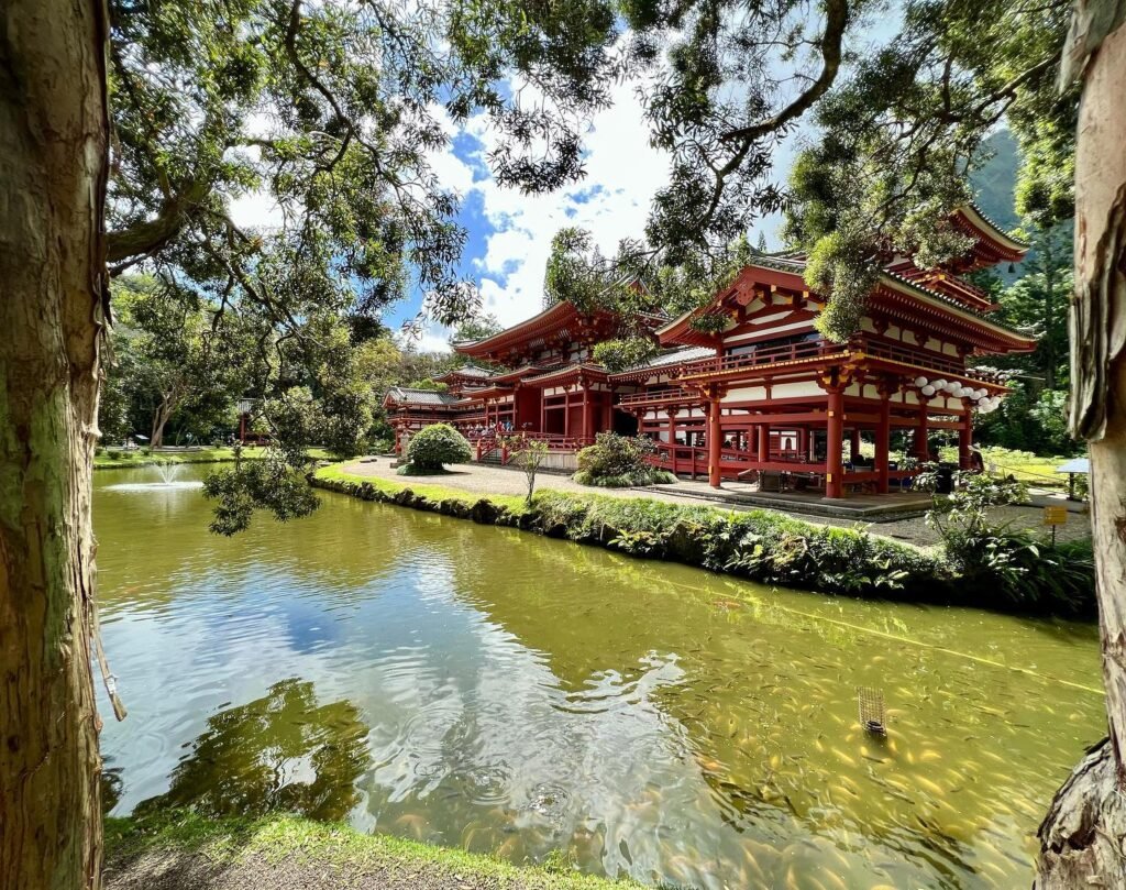 A serene Japanese garden featuring a red traditional building beside a tranquil pond surrounded by tall trees.