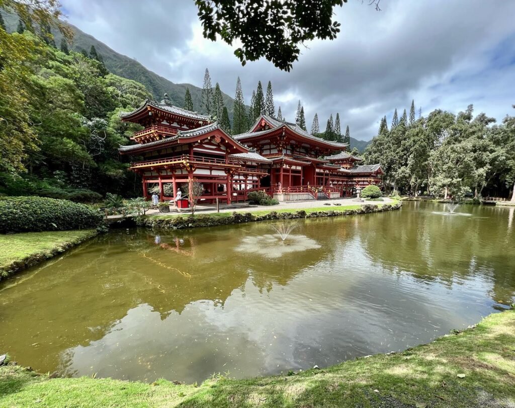 A red Japanese temple complex reflected in a pond, set against a backdrop of mountains and trees.