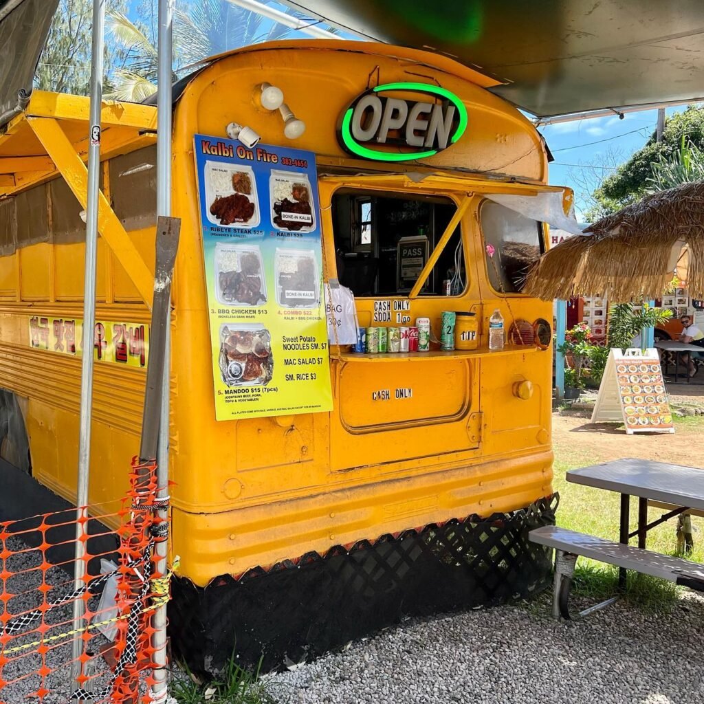 A bright yellow school bus food truck with an 'Open' sign, parked under a canopy with a thatched roof structure in the background.
