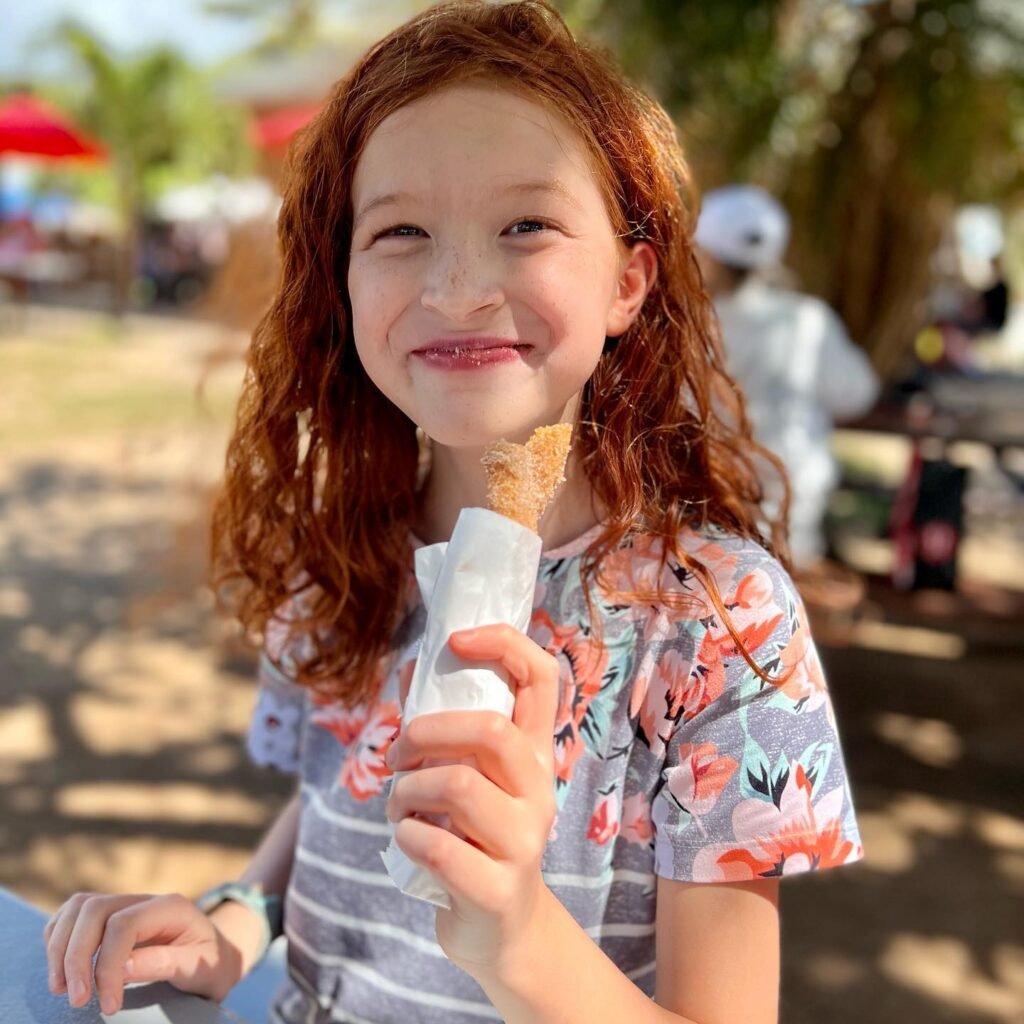 A child with red hair smiles while holding an ice cream cone outside. The background features blurred beach umbrellas and palm trees.