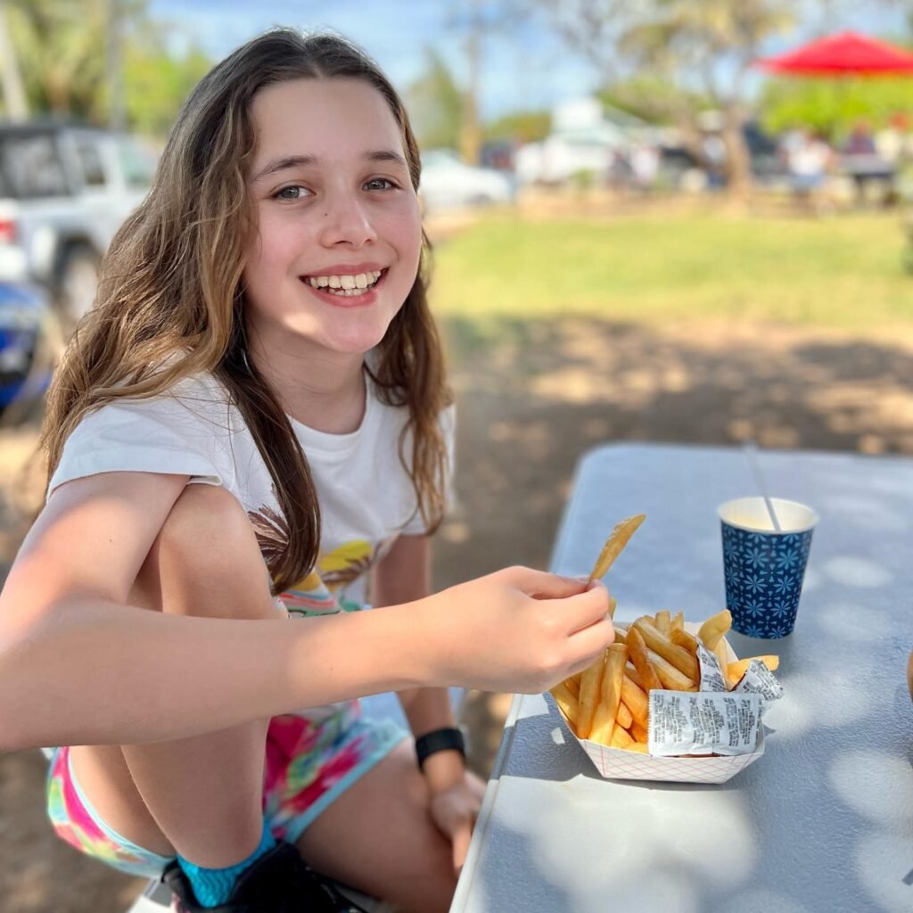 A young person smiles while sitting at an outdoor picnicking table, enjoying french fries and a drink on a sunny day.
