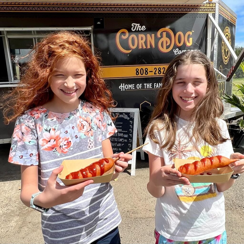 Two smiling children holding fresh corn dogs in front of The Corn Dog food truck.