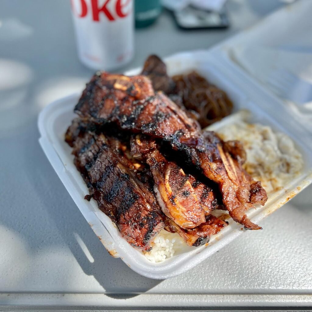 A plastic container filled with grilled ribs and white rice, with a bottle of soda in the background.
