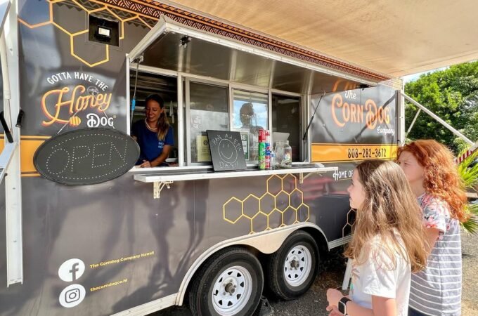 A food truck with 'Hoggy Dog' branding and a 'Cornbox' banner serves customers under a shade structure.