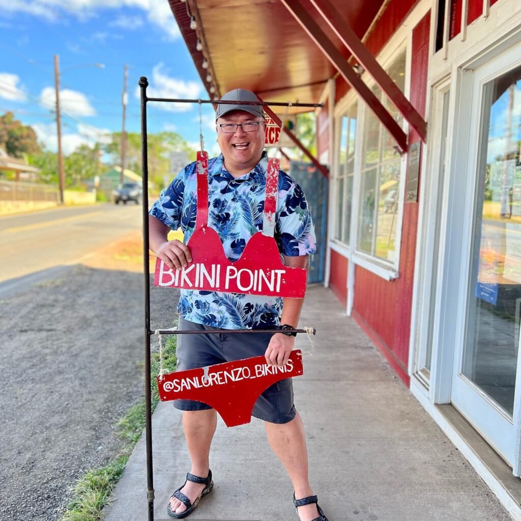 A visitor poses with a red bikini-shaped prop labeled 'Bikini Point' and '@SANLORENZO' outside a building on a sunny day.