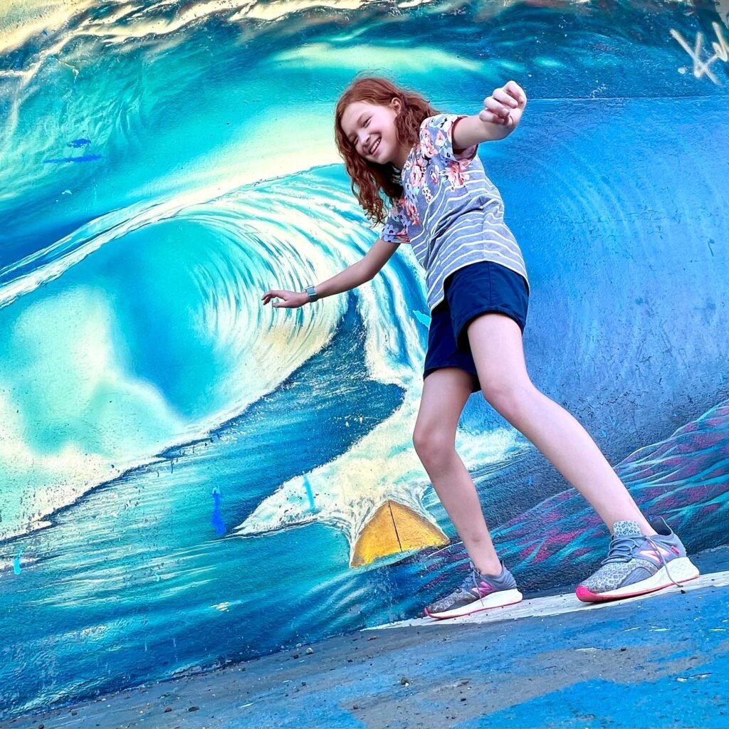 A person strikes a fun surfing pose in front of a vivid mural of a massive ocean wave.