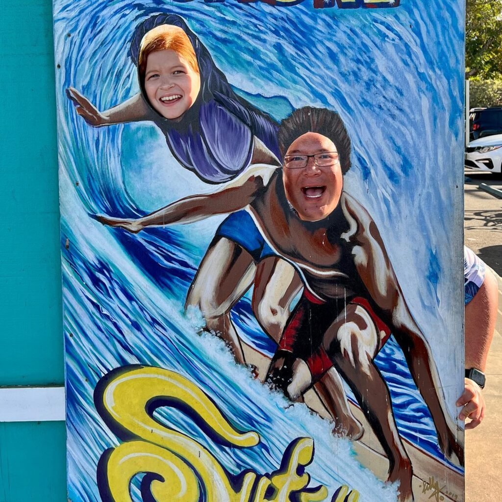 A fun, painted surfboard display featuring cartoon-like portraits of two people surfing, with words in a stylized font below the image.