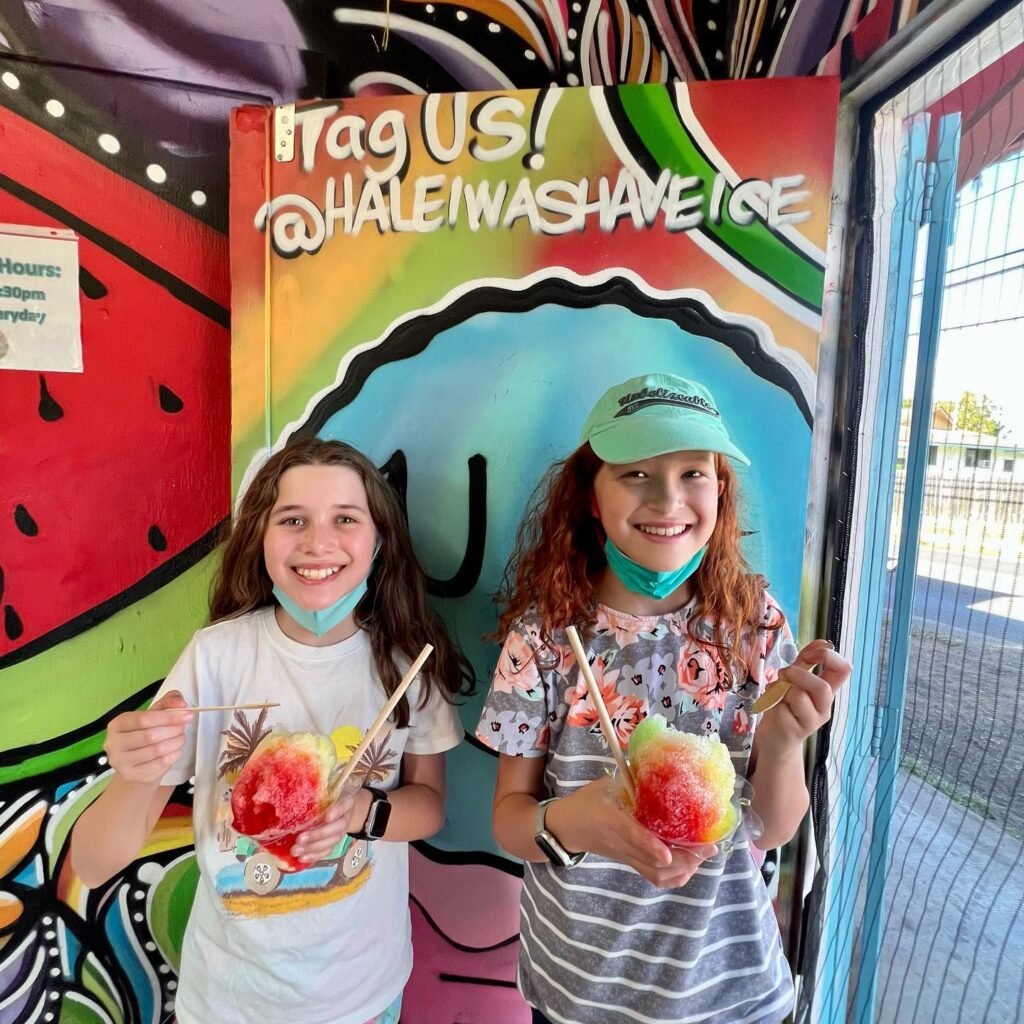 Two people holding colorful shaved ice cones in front of a vibrant mural with the text 'Tag Us! @Haleiwashaveice'.