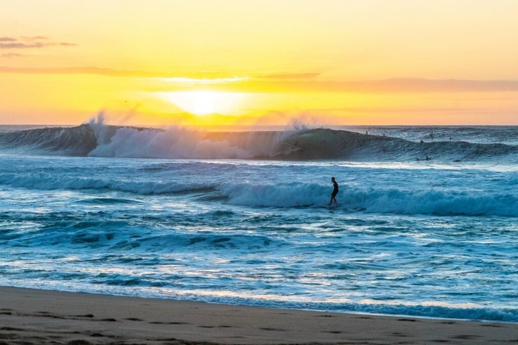 A stunning sunrise over the ocean with waves crashing and a lone surfer silhouetted in the water. The image captures the vibrant colors of the morning sky reflecting on the blue sea.