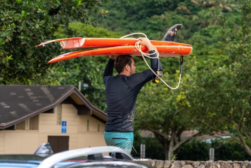 A person in a black top and teal shorts lifts an orange kayak above their head, with a small building, parked cars, and lush green trees in the background.