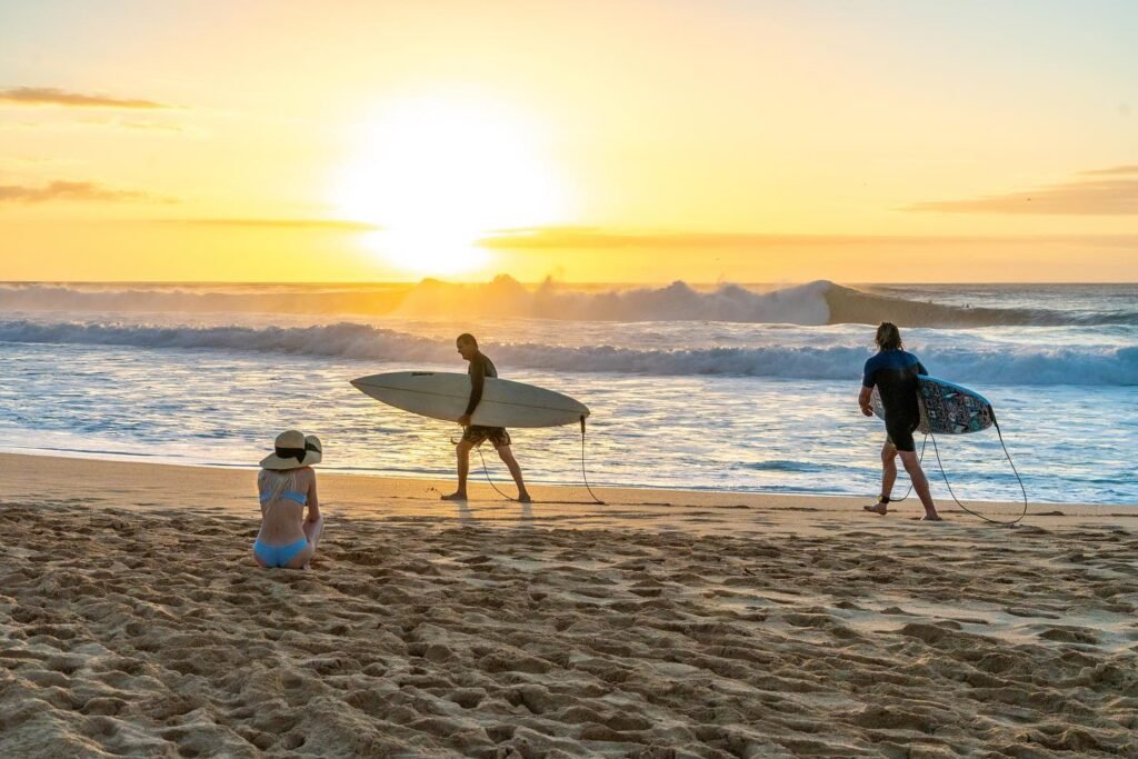 A person carrying a surfboard walks into the ocean at sunset. The sky is golden with gentle waves in the foreground.