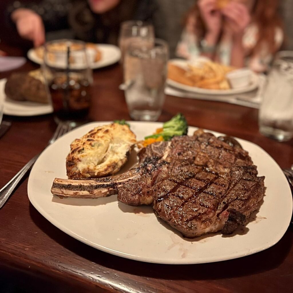 A grilled ribeye steak with visible grill marks and a roasted potato served on a white plate at a dinner table. Glasses of water and a soft drink are in the background with other diners and plates.