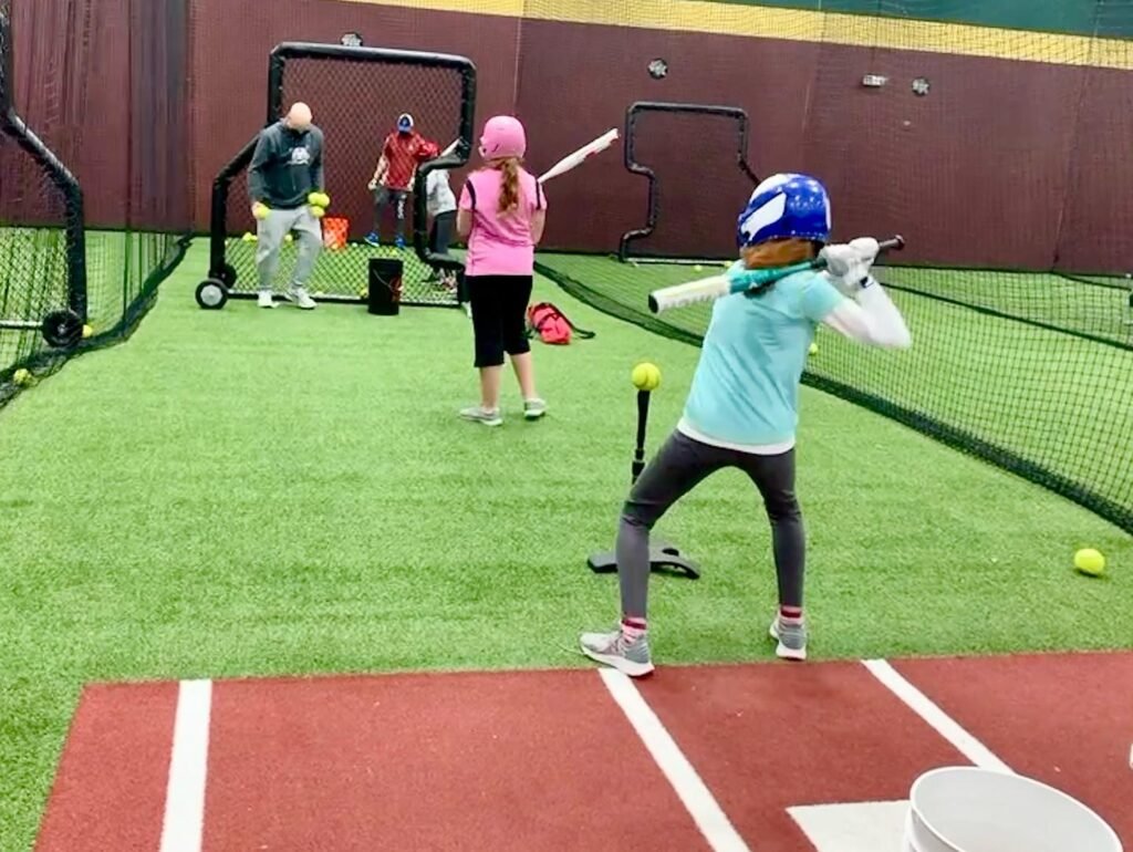 A young teacher in a blue helmet practices a tee ball swing at an indoor sports facility while other children and adults are in the background.