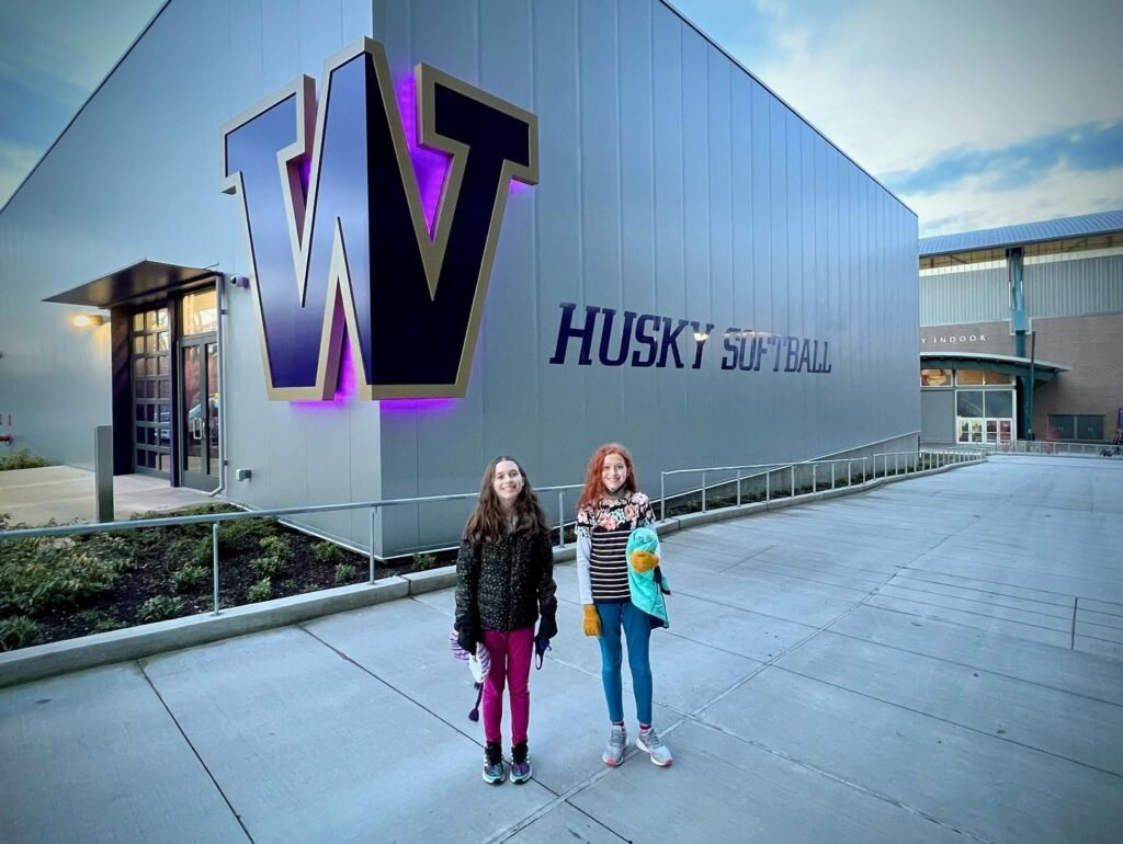 Two visitors posing in front of Husky Softball stadium. Close-up of the large 'W' logo on the building.