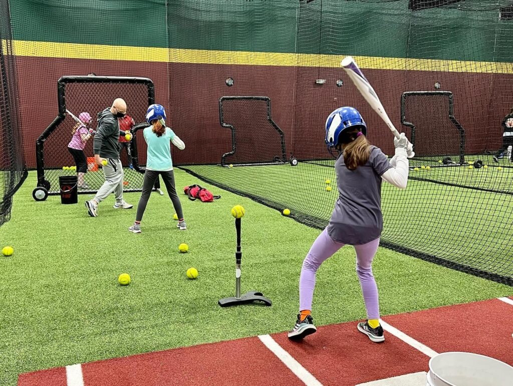 Children wearing helmets and holding bats practice softball in an indoor court with batting nets.