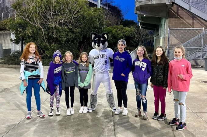 A group of young athletes poses with a raccoon mascot in front of outdoor bleachers under a clear blue sky.