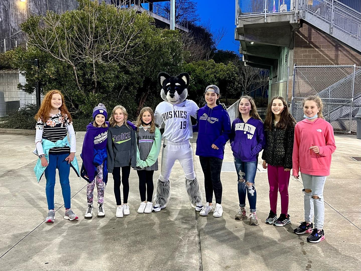 Fastpitch A group of young athletes poses with a raccoon mascot in front of outdoor bleachers under a clear blue sky.