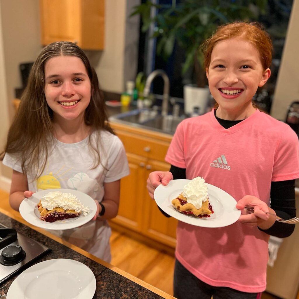 Two smiling children hold plates of apple pie with whipped cream in a warm, home kitchen.