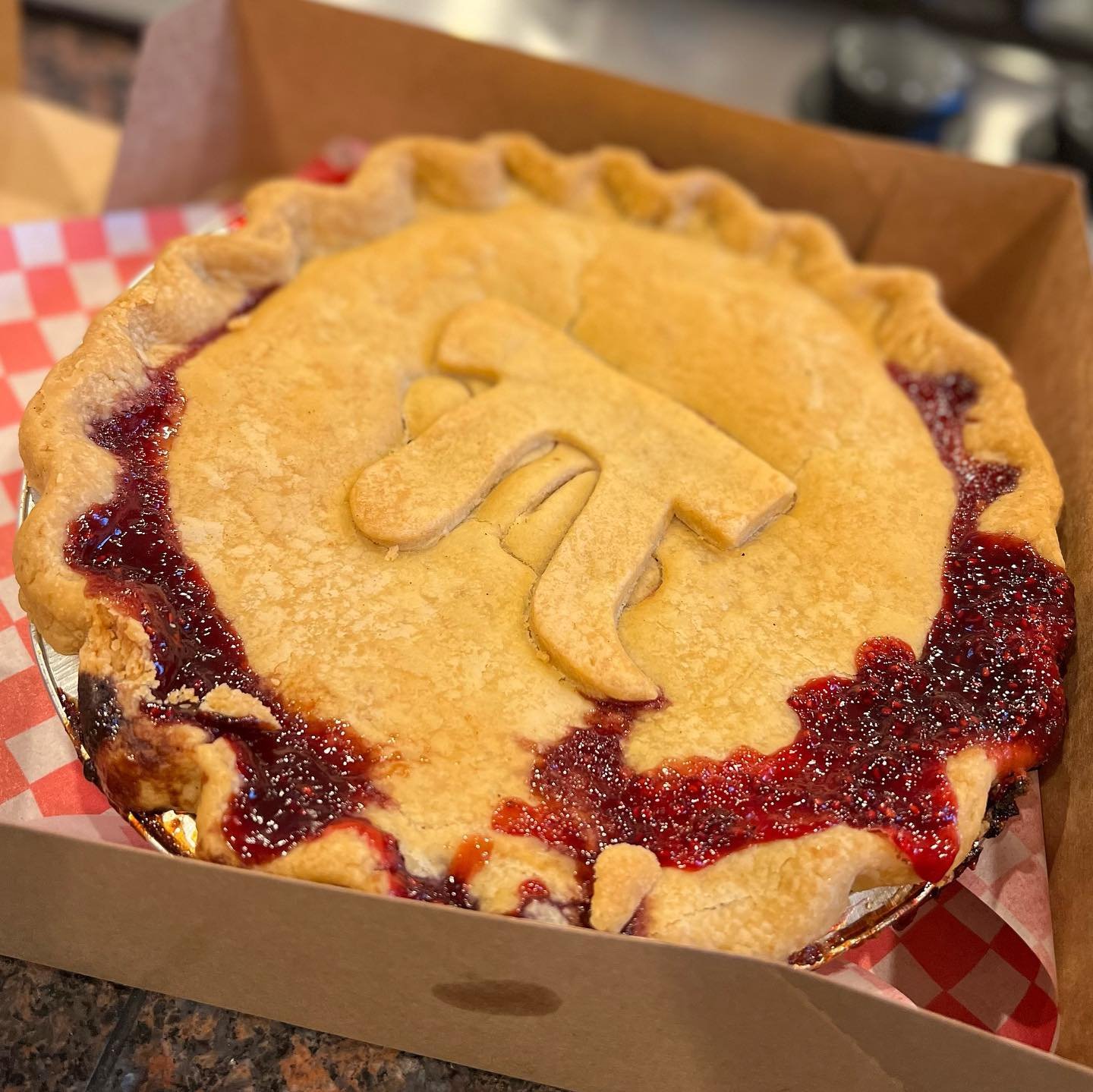 Happy Pi Day Two photos of a rustic cherry pie in a cardboard box lined with red checkered paper, featuring a golden crust with a decorative cut-in design.