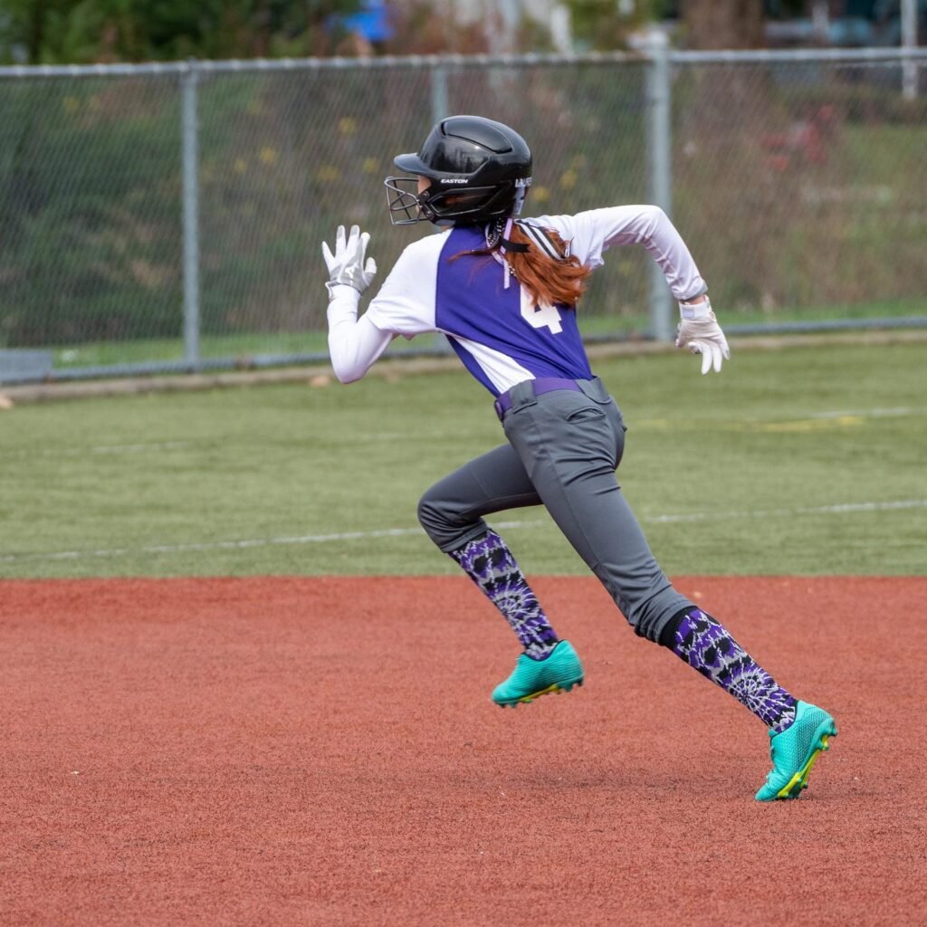 A softball player in a blue jersey and bright green cleats runs on a dirt infield.