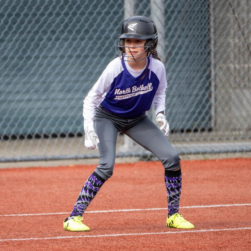 A young athlete in a blue Marist softball uniform crouches on the red field wearing a protective helmet and gloves. The background shows a silver chain-link fence behind them.