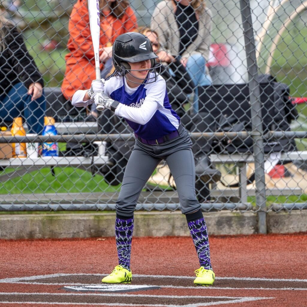 A young softball player standing at home plate, holding a bat, wearing a batter’s helmet and bright socks.