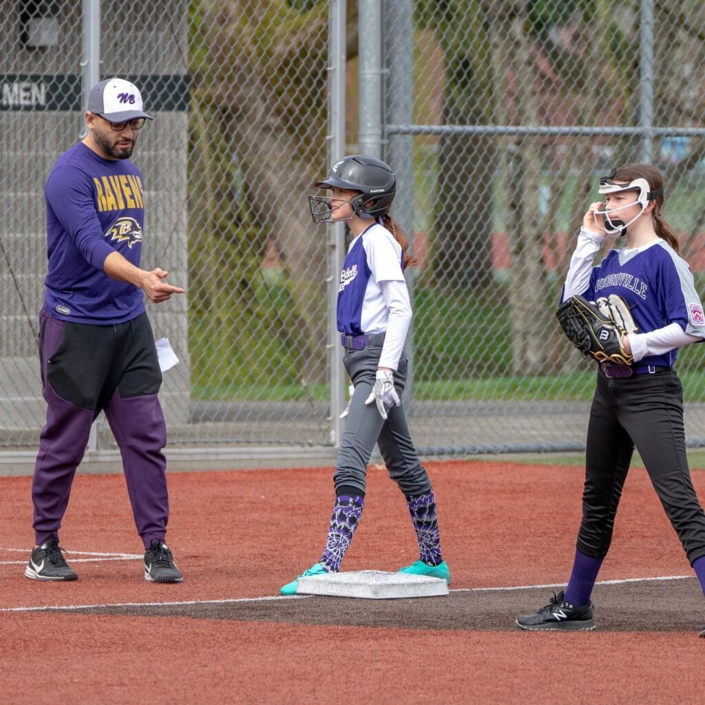 A coach in purple team gear pointing to a base while two players stand nearby on the field. The scene captures a moment of instruction during a youth softball game.