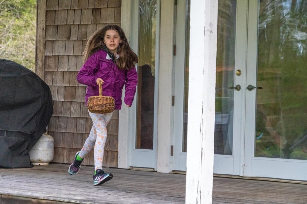 A child carrying a basket walks across a wooden porch next to a white door and cedar shingles.