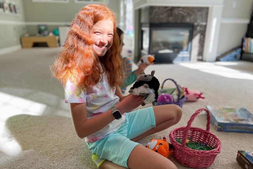 A child with long red hair sits on the floor holding stuffed animals near a pink Easter basket.