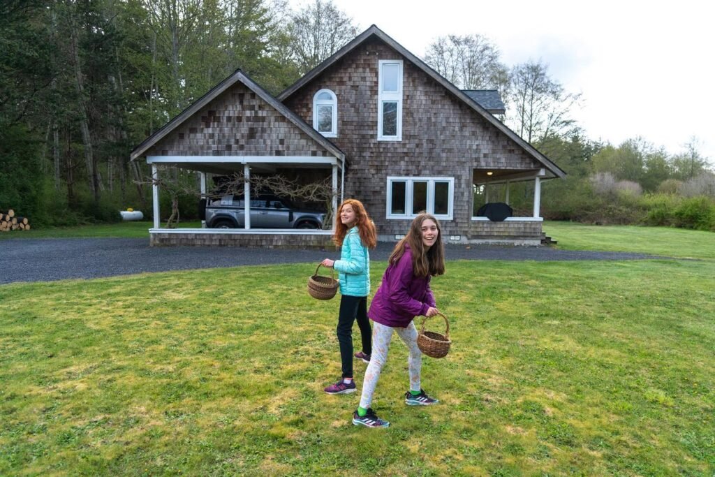 Children holding baskets explore a grassy lawn in front of a rustic stone house surrounded by tall evergreen trees.