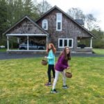 Orca Watching Children holding baskets explore a grassy lawn in front of a rustic stone house surrounded by tall evergreen trees.