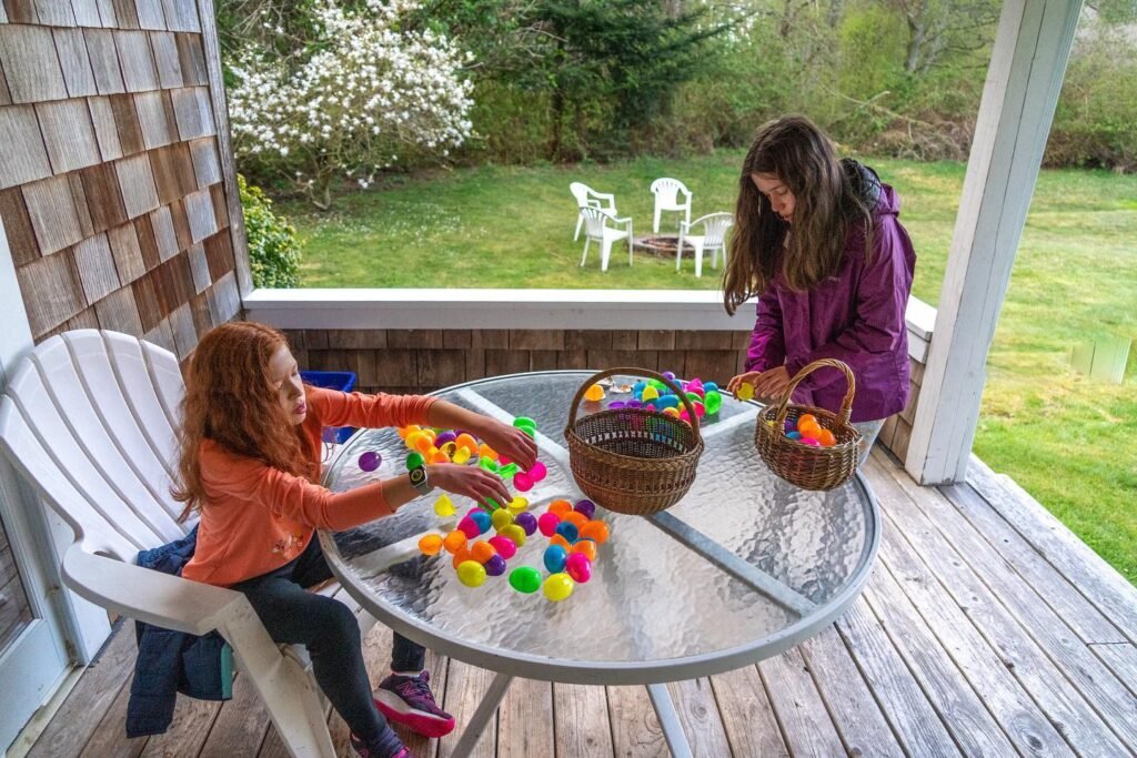 A child and an adult sort colorful plastic eggs into baskets on a wooden porch table. A lush green garden is visible in the background.